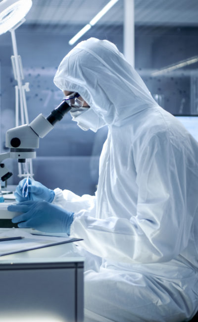 In a Secure High Level Laboratory Scientists in a Coverall Conducting a Research. Biologist Adjusts Samples in a  Petri Dish with Pincers and Examines Them Under Microscope and His Colleague Analyzes Results on a Computer.